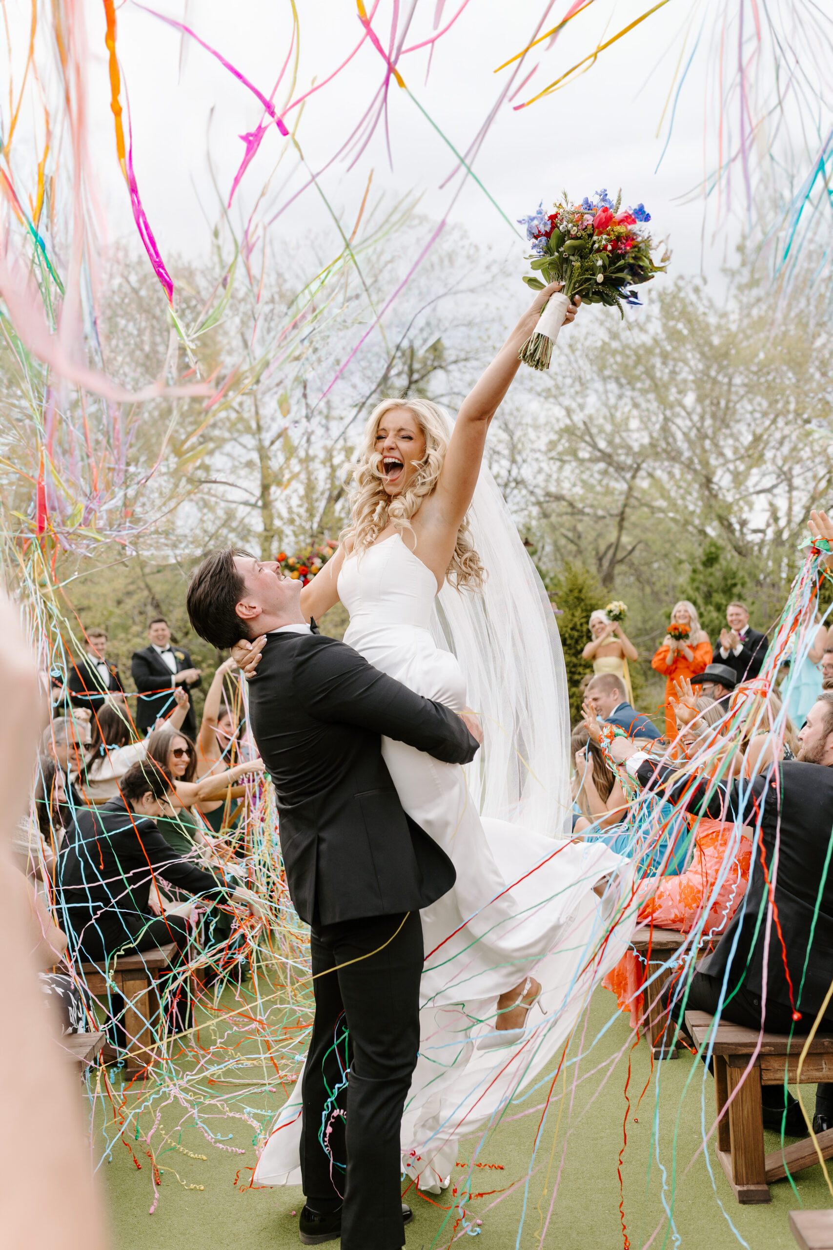 Groom lifting bride joyfully at the end of their wedding ceremony with colorful confetti flying in the air at The Barn at Timber Point.