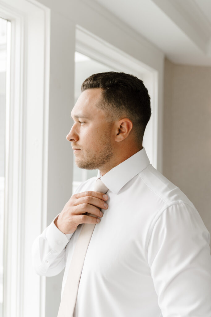 groom adjusts tie looking out window on wedding day