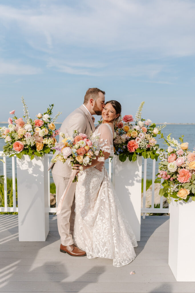 groom kisses bride on cheek with pastel florals behind them