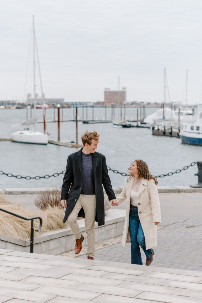 Couple walking along East Boston waterfront engagement session with harbor views and sailboats