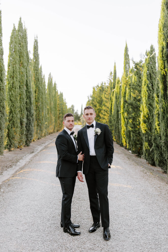 grooms look at camera holding hands in tuscany with tall trees on each side