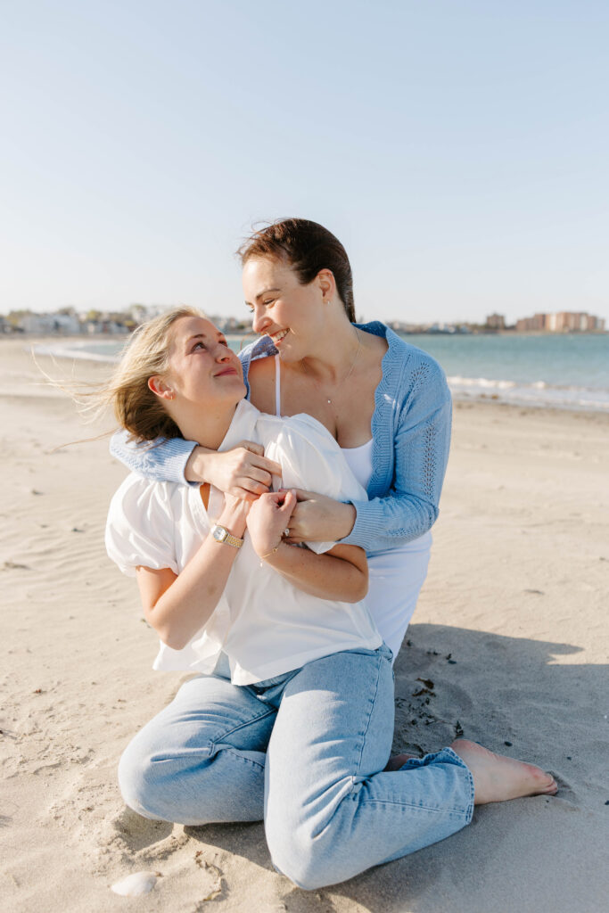 Couple sitting on sand in East Boston looking at each other laughing