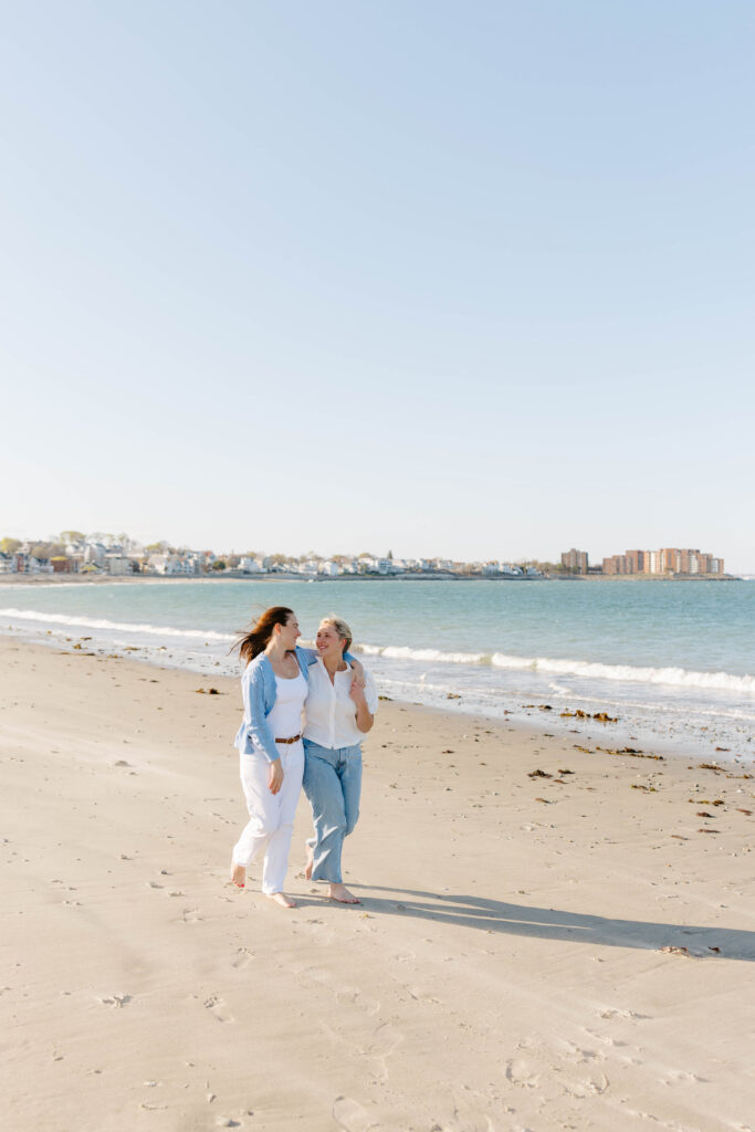Couple walking barefoot along East Boston beach with ocean behind them