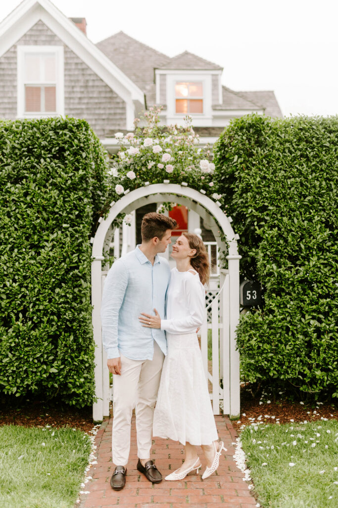 couple smiles at each other with green shrubs in the background with white fence