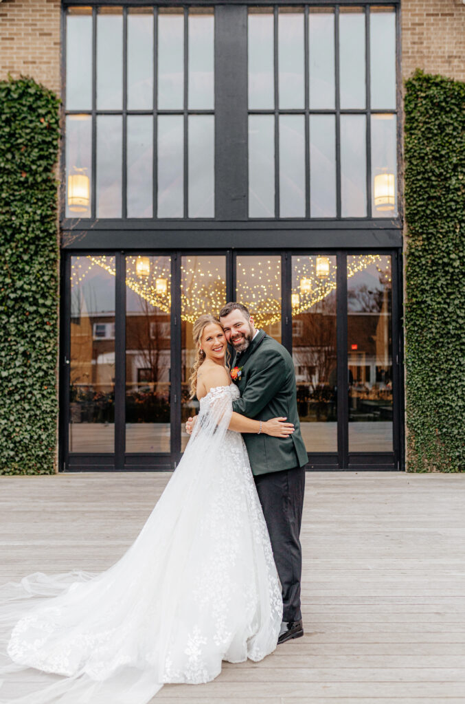 bride and groom smile at camera in front of greenery building