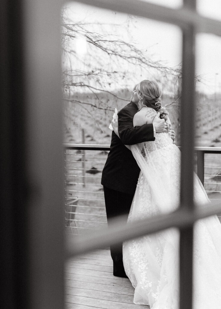 Black and white photo looking through window of father and daughter hug on wedding day