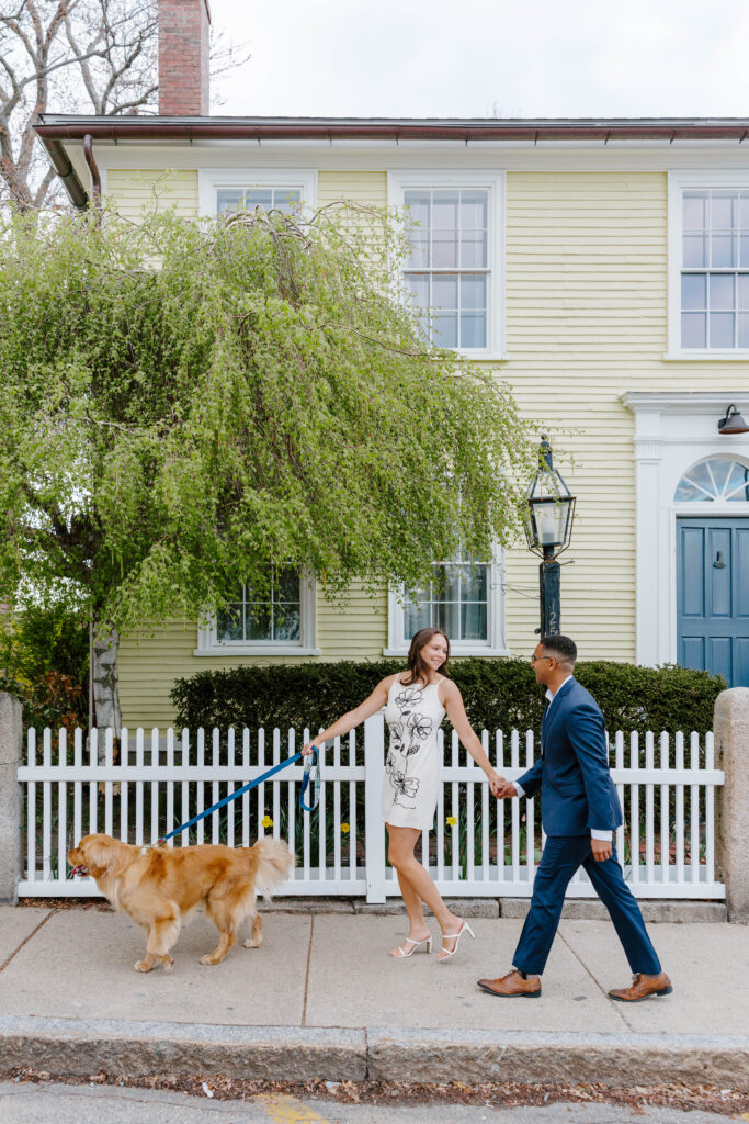 Couple walking their golden retriever along a sidewalk in downtown Rockport Massachusetts, holding hands in front of a charming yellow coastal home with white picket fence and spring greenery