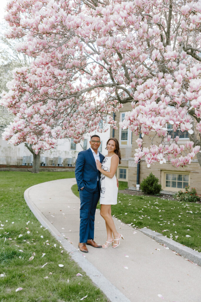 Couple standing together beneath blooming magnolia trees in Rockport Massachusetts, smiling at the camera along a curved walkway with soft pink petals scattered on the ground
