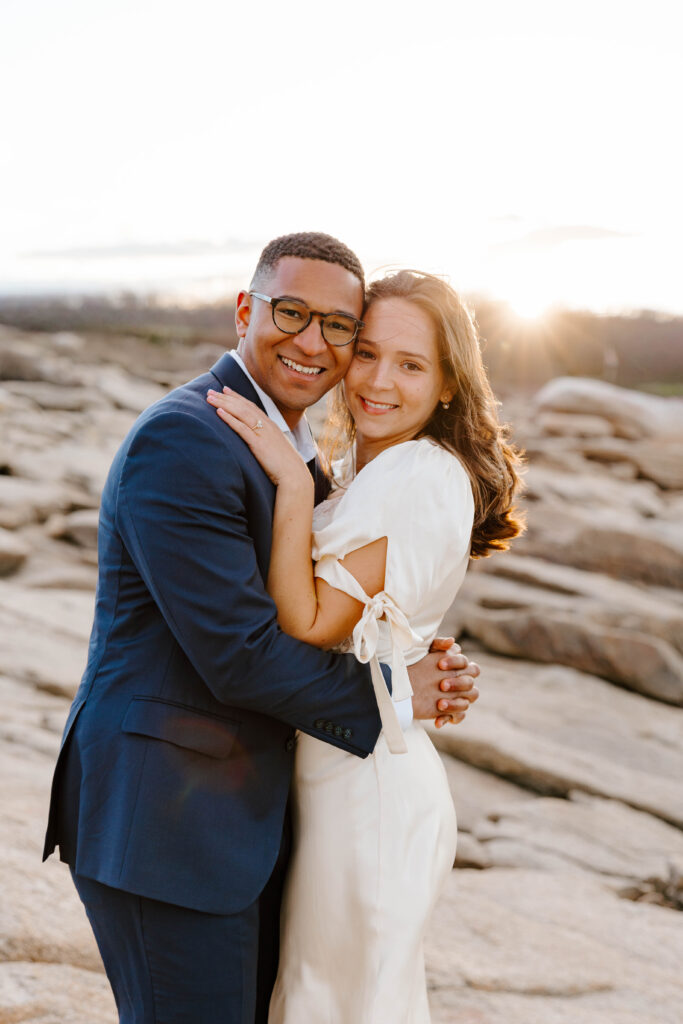 Couple embracing during golden hour on a rocky beach with warm sunset light and ocean in the background
