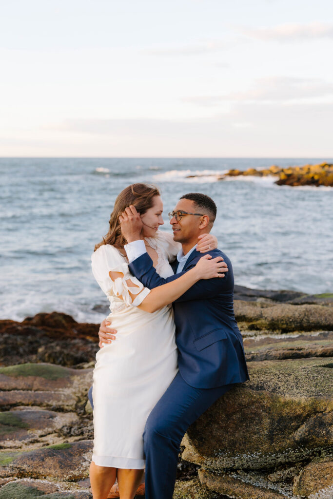 Couple embracing on rocky coastline in Rockport Massachusetts at sunset, smiling at each other with ocean waves and soft pastel sky in the background