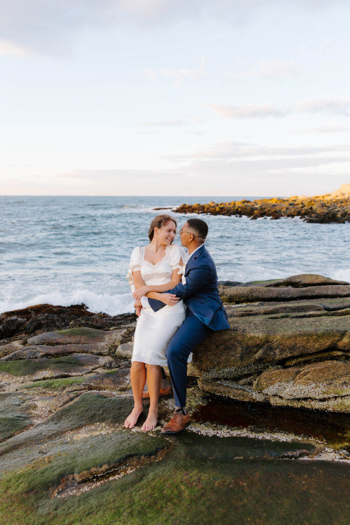 Couple sitting on seaweed covered rocks along the Rockport Massachusetts shoreline at sunset, sharing a quiet moment with waves crashing behind them