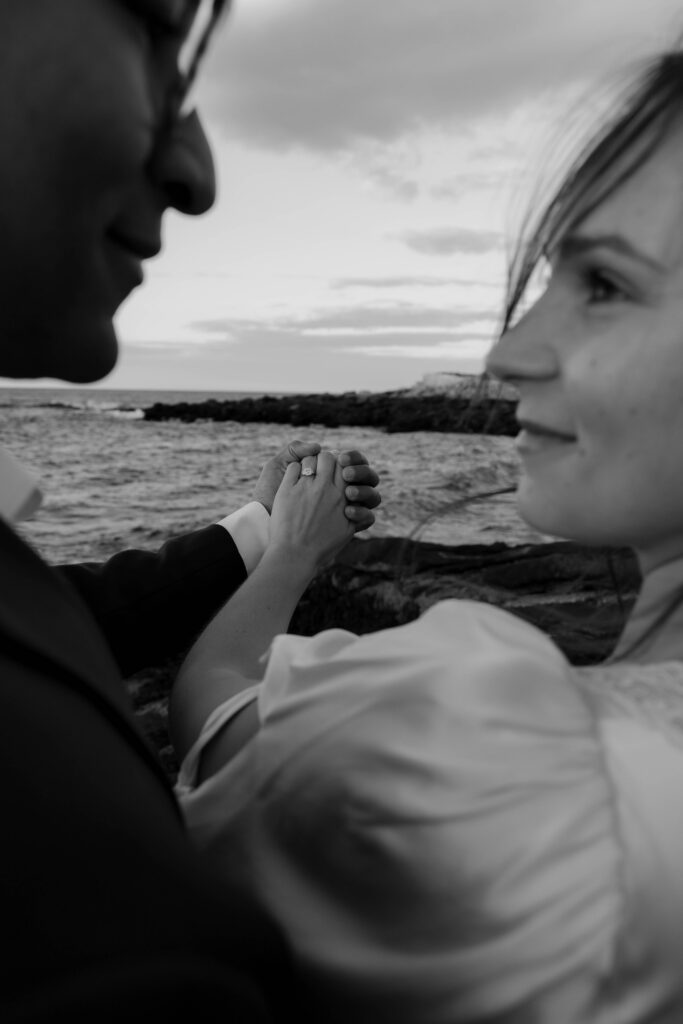 Close up black and white photo of couple holding hands with engagement ring visible, ocean and rocky coastline in background in Rockport Massachusetts