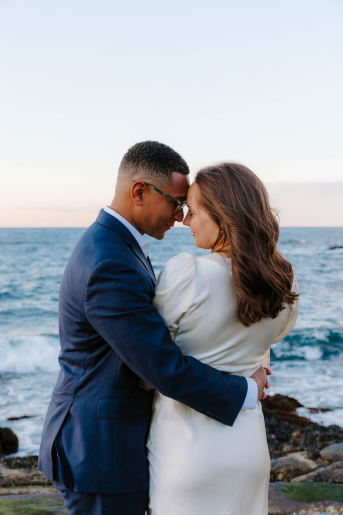 Couple embracing at sunset along the rocky coastline in Rockport Massachusetts with ocean waves in the background