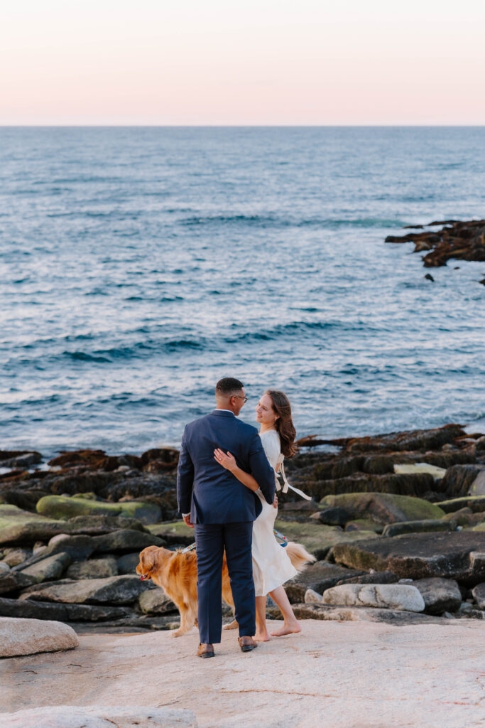 Couple walking along rocky shoreline in Rockport Massachusetts at sunset with their golden retriever and ocean views behind them