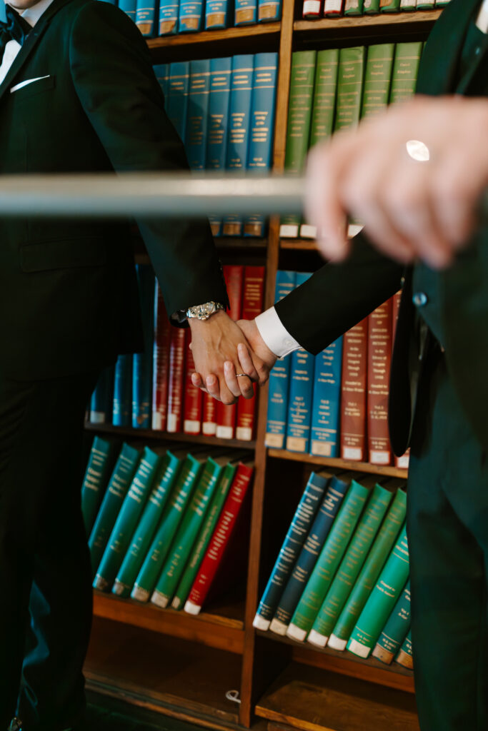 Close up detail photo of Ryan and Spencer holding hands in front of colorful bookshelves at the Boston Public Library.