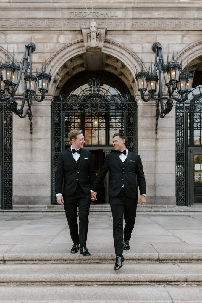 Ryan and Spencer walking hand in hand outside the Boston Public Library in black tuxedos before their wedding ceremony.