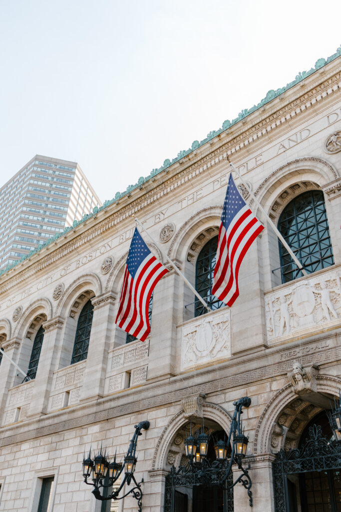Exterior of the Boston Public Library in Boston with American flags hanging above the entrance on a bright summer day.