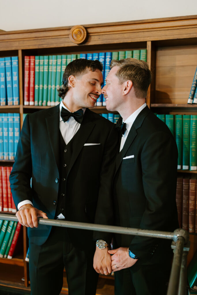 Romantic portrait of Ryan and Spencer standing together in front of colorful bookshelves inside the Boston Public Library.