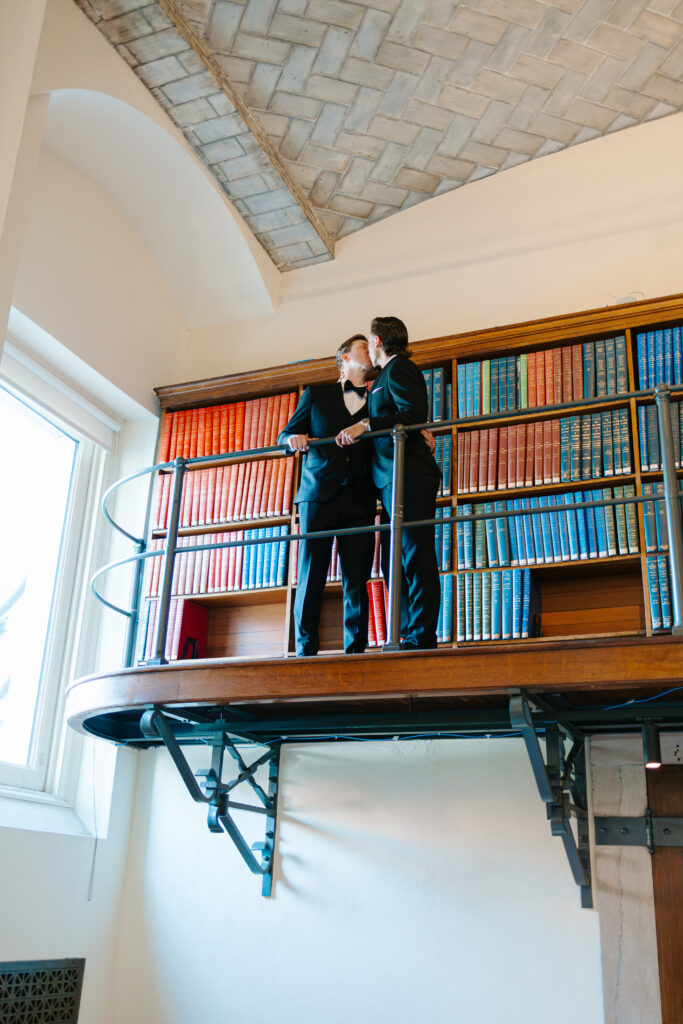 Ryan and Spencer kissing on the upper library balcony surrounded by colorful bookshelves inside the Boston Public Library.