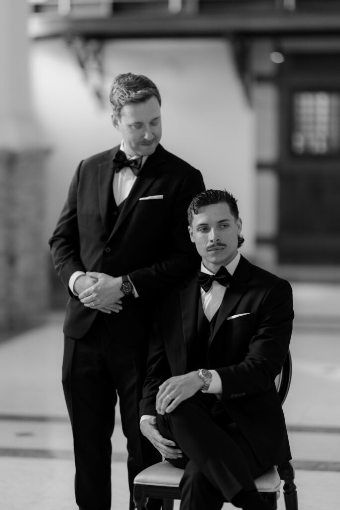 Editorial black and white portrait of Ryan standing beside Spencer seated in a chair during their Boston wedding portraits.