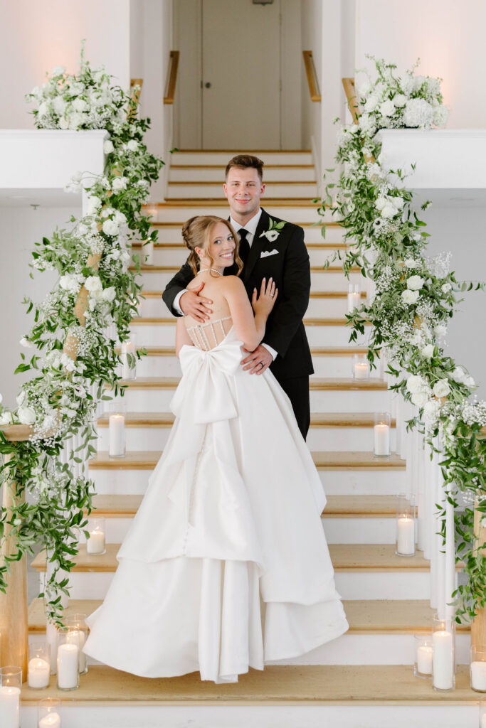 Bride and groom smile at camera at Belle Mer on staircase with green and white floral on the railings