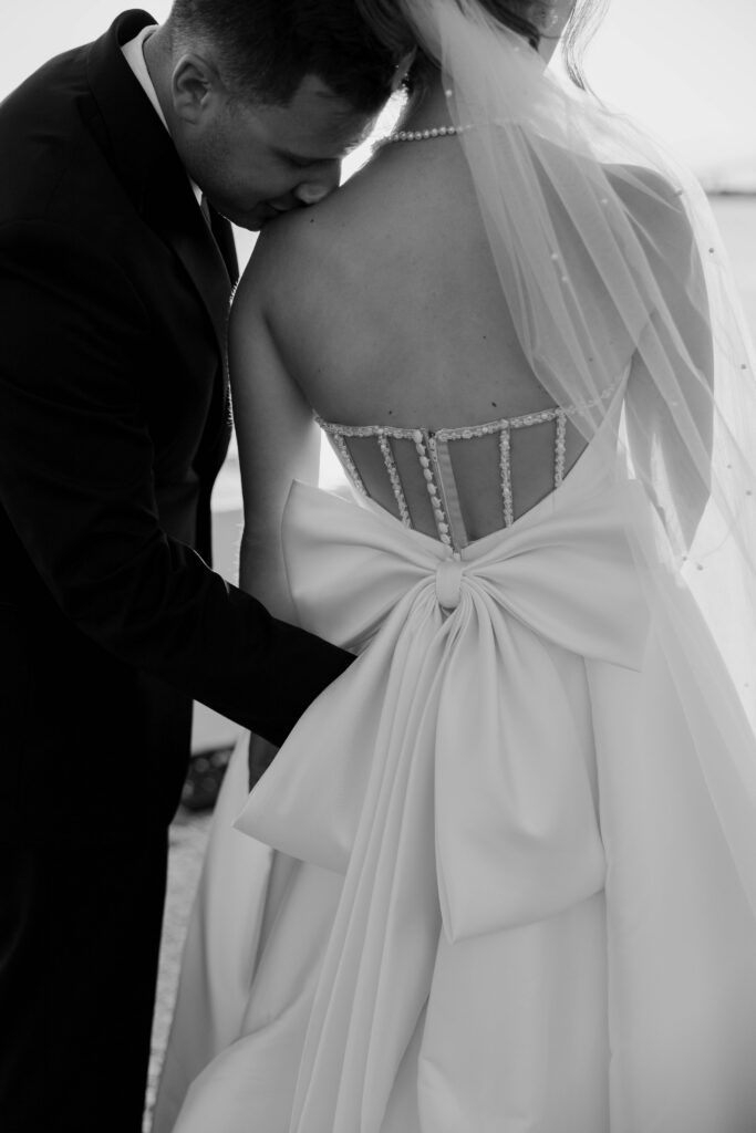Black and white wedding portrait of groom kissing brides shoulder with back of wedding dress and a bow