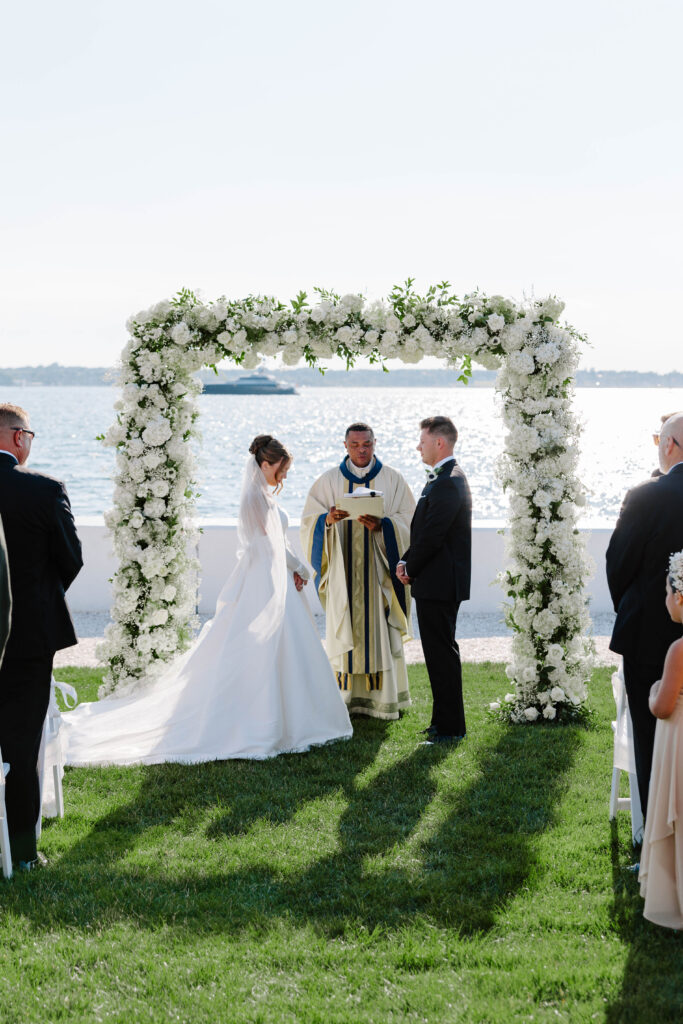 Bride and groom during ceremony with floral arbor in front of ocean in Newport