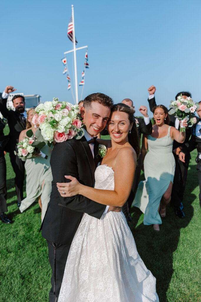 bride and groom embrace with wedding party celebrating behind them