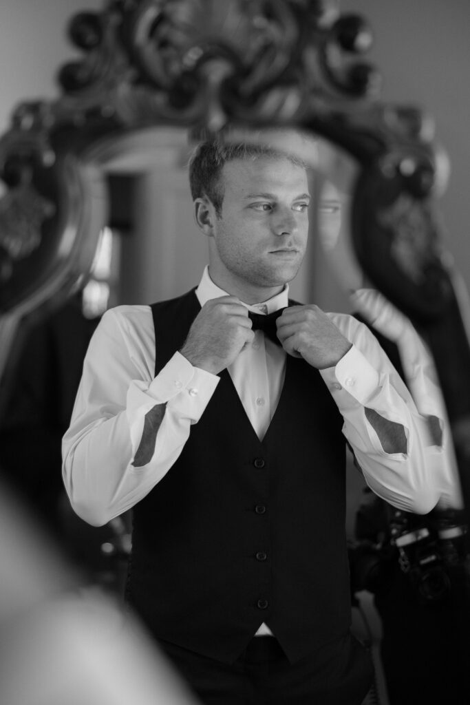 Black and white photo of groom tying tie in front of mirror